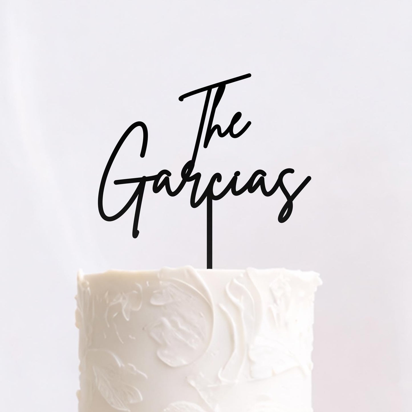Black acrylic script wedding cake topper reading “The Garcias” displayed on textured white wedding cake. Made by Honey Craft Studios