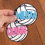 A hand holds two personalized volleyball-shaped tags with the names 'WILLOW' in light blue and 'QUINN' in pink, set against a wooden background.