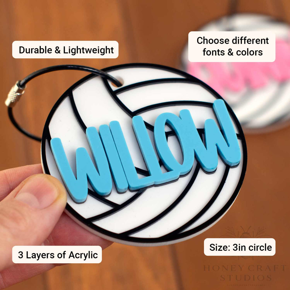 A hand holds a round acrylic name plaque shaped like a volleyball, with the name 'WILLOW' raised in bright blue letters. The image includes text callouts reading 'Durable & Lightweight,' 'Choose different fonts & colors,' '3 Layers of Acrylic,' and 'Size: 3in circle,' on a warm wood background.